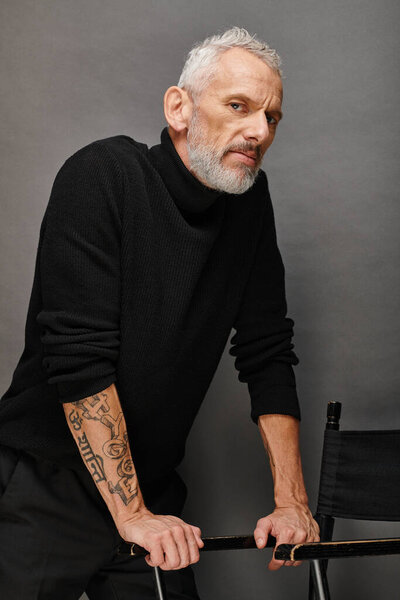 fashionable handsome mature man in chic turtleneck posing net to folding chair and looking at camera