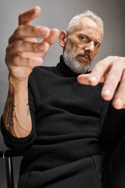 stock image handsome mature man with beard in stylish turtleneck sitting on folding chair and looking at camera