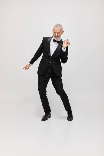 appealing merry mature male model with gray beard and bow tie in elegant tuxedo dancing actively