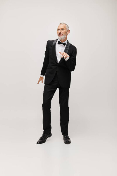 хорошо одетый зрелый мужчина модель с бабочкой галстук в шикарный черный смокинг танцы активно на сером фоне