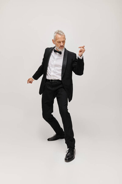 good looking mature male model with bow tie and beard in elegant tuxedo dancing on gray backdrop