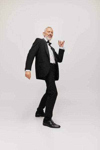 cheerful attractive gray bearded man with dapper stylish tuxedo dancing and smiling at camera
