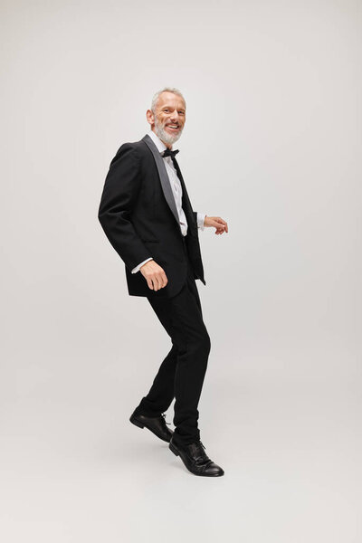 cheerful attractive gray bearded man with dapper stylish tuxedo dancing and smiling at camera