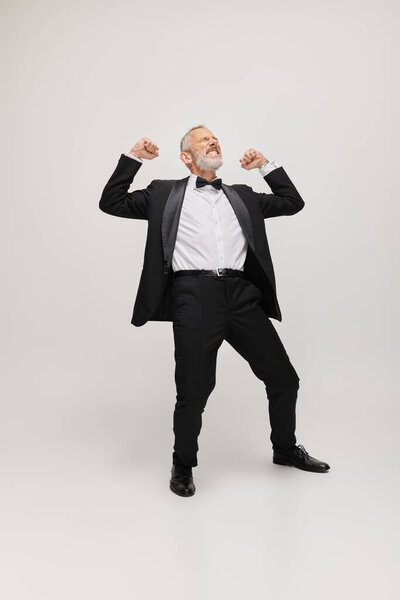 joyous good looking mature man in elegant suit with bow tie dancing happily on gray backdrop