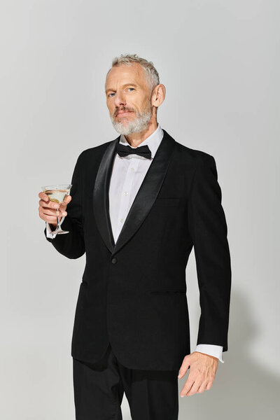handsome bearded mature male model  in elegant tuxedo holding martini glass and looking at camera
