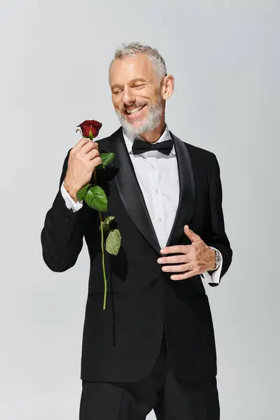 赤いバラを保持し 幸せに笑顔でエレガントなタキシードのひげを持つ魅力的な楽しい成熟した男 — ストック写真