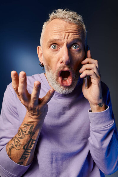 handsome fashionable mature man in purple stylish sweatshirt grimacing while talking by phone