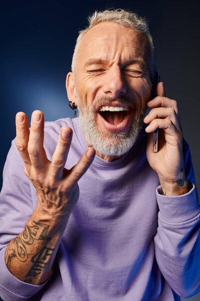 cheerful bearded trendy mature man in purple stylish sweatshirt talking by phone on blue backdrop