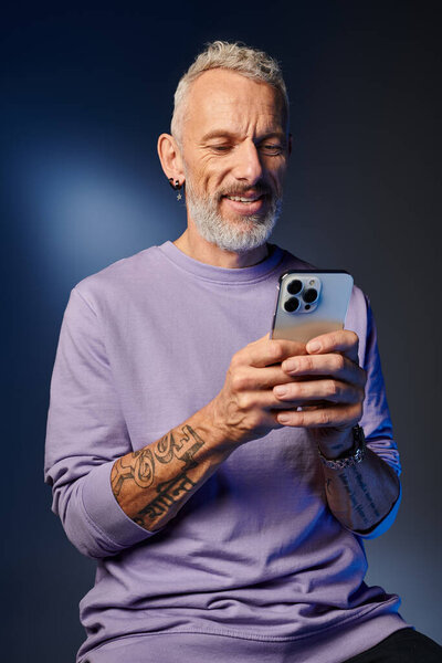 joyous good looking mature man in purple sweatshirt with gray beard looking at his smartphone