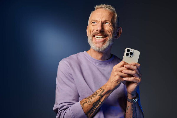 good looking joyous mature man with beard in fashionable sweatshirt holding phone and looking away