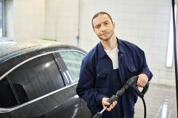cheerful handsome serviceman in uniform holding hose preparing to wash car and smiling at camera