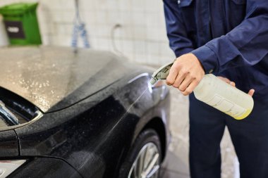 cropped view of devoted serviceman using pulverizer with water to clean black modern car in garage clipart