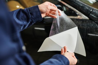 cropped view of hard working man in blue uniform ready to apply protective foil on black car clipart