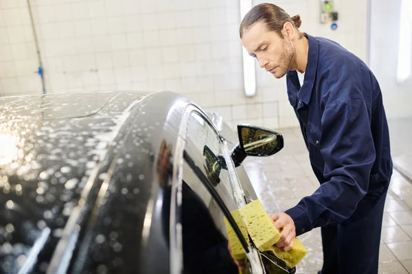 appealing hard working serviceman in blue uniform washing car with soapy sponge while in garage