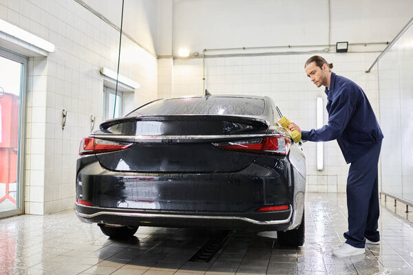 handsome dedicated professional worker in blue uniform using soapy sponge to clean black car