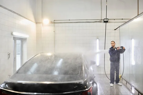 stock image good looking devoted serviceman in blue uniform with collected hair using hose to wash black car