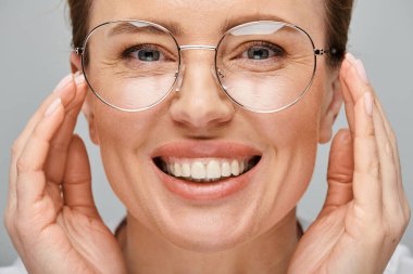 joyous beautiful woman with blonde collected hair and glasses smiling at camera on gray backdrop clipart