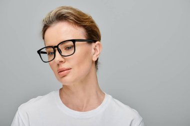 good looking blonde woman with collected hair and glasses posing on gray backdrop and looking away clipart