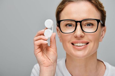 joyful appealing woman with blonde hair and glasses holding contact lenses and looking at camera clipart