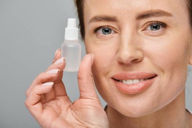 cheerful good looking woman holding eye drops and looking straight at camera on gray backdrop clipart