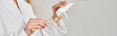 cropped view of adult woman in comfy bathrobe putting tooth paste on her brush near mirror, banner clipart