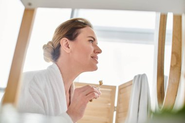 Bornozlu, sarı saçlı, dişlerini fırçalayan ve aynaya bakan neşeli bir kadın.