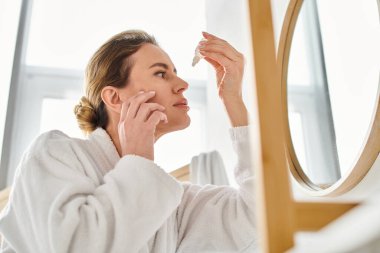 Banyosuna göz damlası damlatan bornozlu, saçları toplanmış sarışın bir kadın.