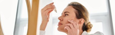 appealing blonde woman with collected hair in bathrobe putting in eye drops in her bathroom, banner clipart