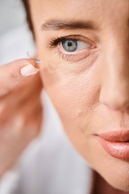 appealing woman in white comfy bathrobe wearing her contact lenses near mirror in bathroom clipart