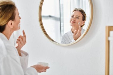 Rahat beyaz bornozlu güzel sarışın kadın evde banyoda yüz kremi sürüyor.