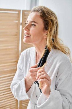 joyous appealing blonde woman in cozy bathrobe using flat iron on her hair while in bathroom clipart