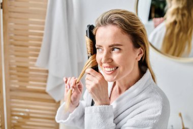 joyous appealing blonde woman in cozy bathrobe using flat iron on her hair while in bathroom clipart