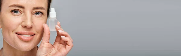 stock image joyous good looking woman holding eye drops and looking straight at camera on gray backdrop, banner