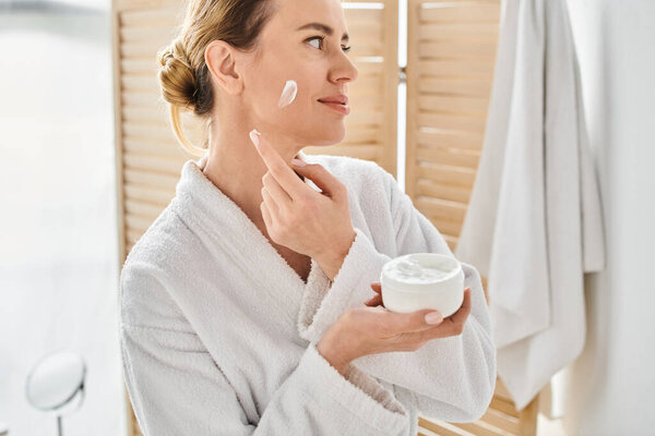 appealing blonde woman in comfy white bathrobe applying face cream while in bathroom at home