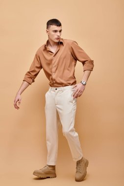 Bej gömlekli, pantolonlu ve botlu bej arkaplanda poz veren genç bir adam.