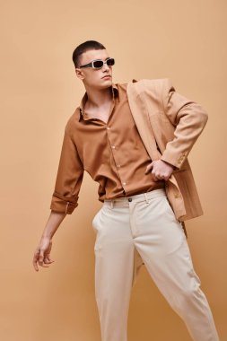 Bej ceketli, omuz, gömlek, pantolon ve bej arka planda güneş gözlüğü takan şık bir adam.