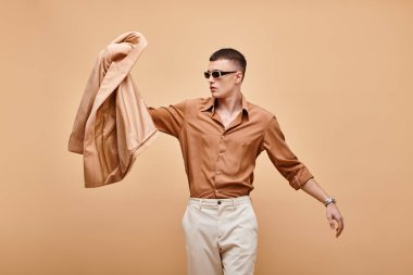 Bej gömlekli, güneş gözlüklü ve elinde bej arka planda ceketi olan bir adamın moda fotoğrafı.
