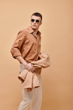 Bej gömlekli, güneş gözlüklü ve elinde bej arka planda ceketi olan bir adamın moda fotoğrafı.