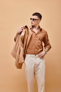 Bej gömlekli, güneş gözlüklü ve elinde bej arkaplanda ceketi olan bir adamın moda fotoğrafı.