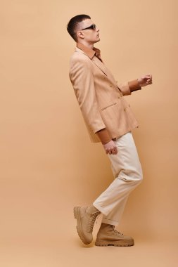 Bej ceketli, gömlekli, pantolonlu ve botlu bej arkaplanda poz veren süslü bir adamın yan görüntüsü.