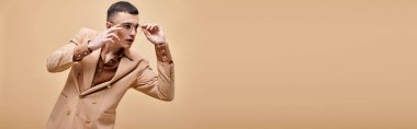 Handsome man in beige jacket touching glasses posing on peachy beige background, banner clipart