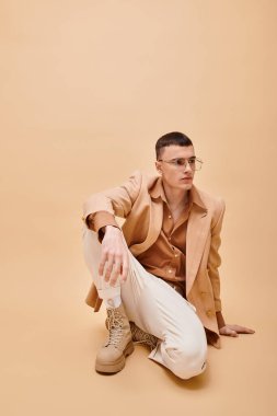 Bej ceketli, gözlüklü, şeftali rengi arka planda oturan yakışıklı bir adam.