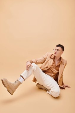 Stylish man in 20s in beige jacket and glasses sitting on peachy beige background clipart