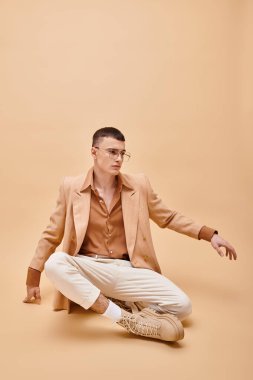 Stylish man in beige jacket and glasses sitting in lotus pose  on peachy beige background clipart