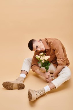 Bej gömlekli, çiçekli, gözlüklü bej arka planda oturan genç adam kameraya bakıyor.