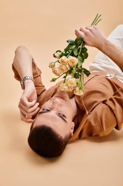 Fashion portrait of man in beige shirt lying with rose flowers bouquet on beige background clipart