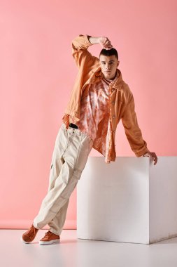 Bej gömlekli, pantalonlu ve botlu, pembe arka planda beyaz küplü, modaya uygun bir adamın resmi.