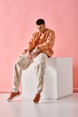 Stylish man in beige shirt, pants and sneakers sitting on white cube on pink background clipart