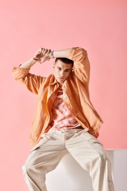 Portrait of man with hands above head  and sitting on white cube on pink background clipart