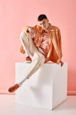 Fashion shot of fashionable man in beige shirt, pants and boots on white cube on pink background clipart